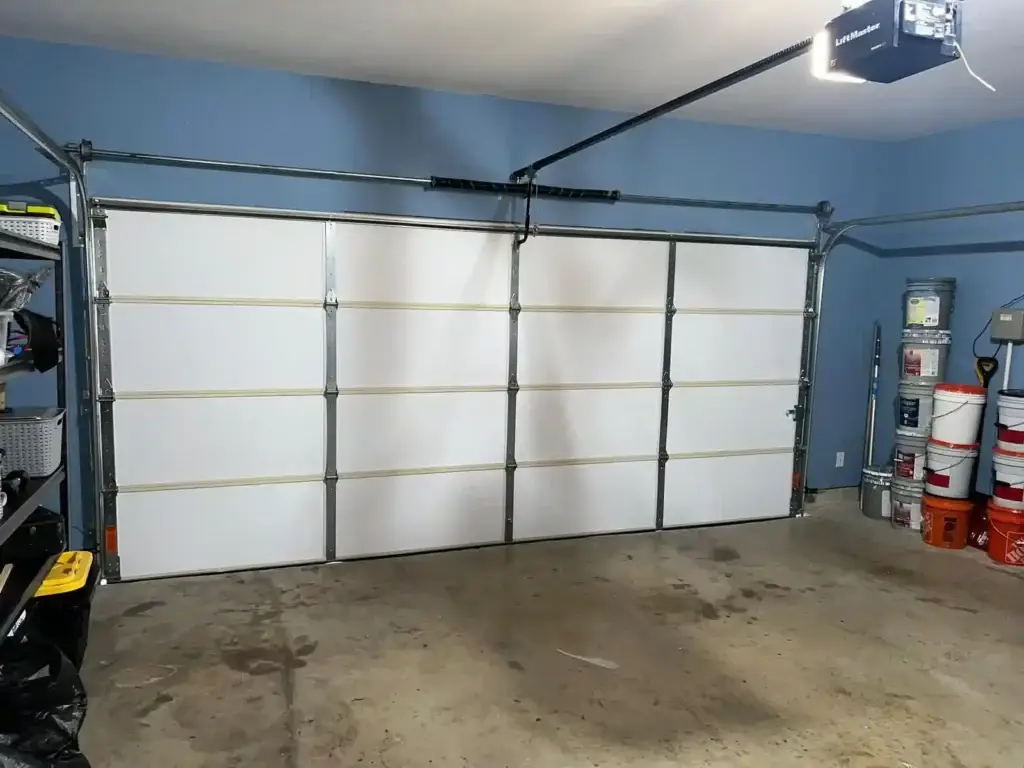 Two-car garage interior with a white sectional door and a ceiling-mounted LiftMaster opener, blue walls, and left/right storage areas with buckets on the right.