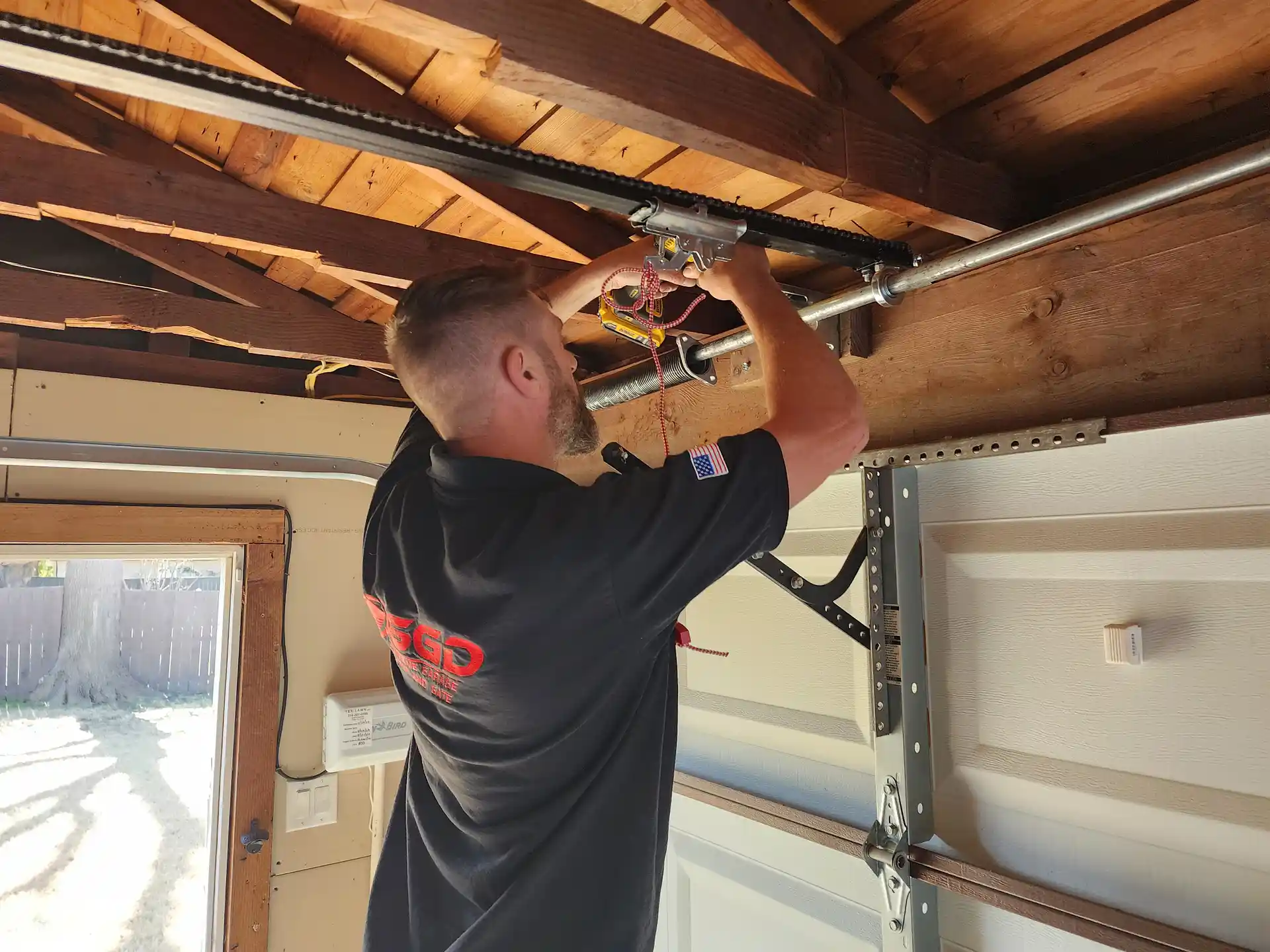 An SGD garage door repair technician adjusting an automatic opener trolley carriage rail beneath an open wood truss ceiling framework.