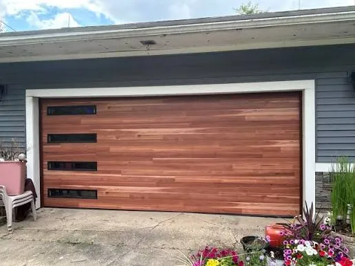 Modern wood-look residential garage door with horizontal panels and narrow black windows on a gray home exterior.